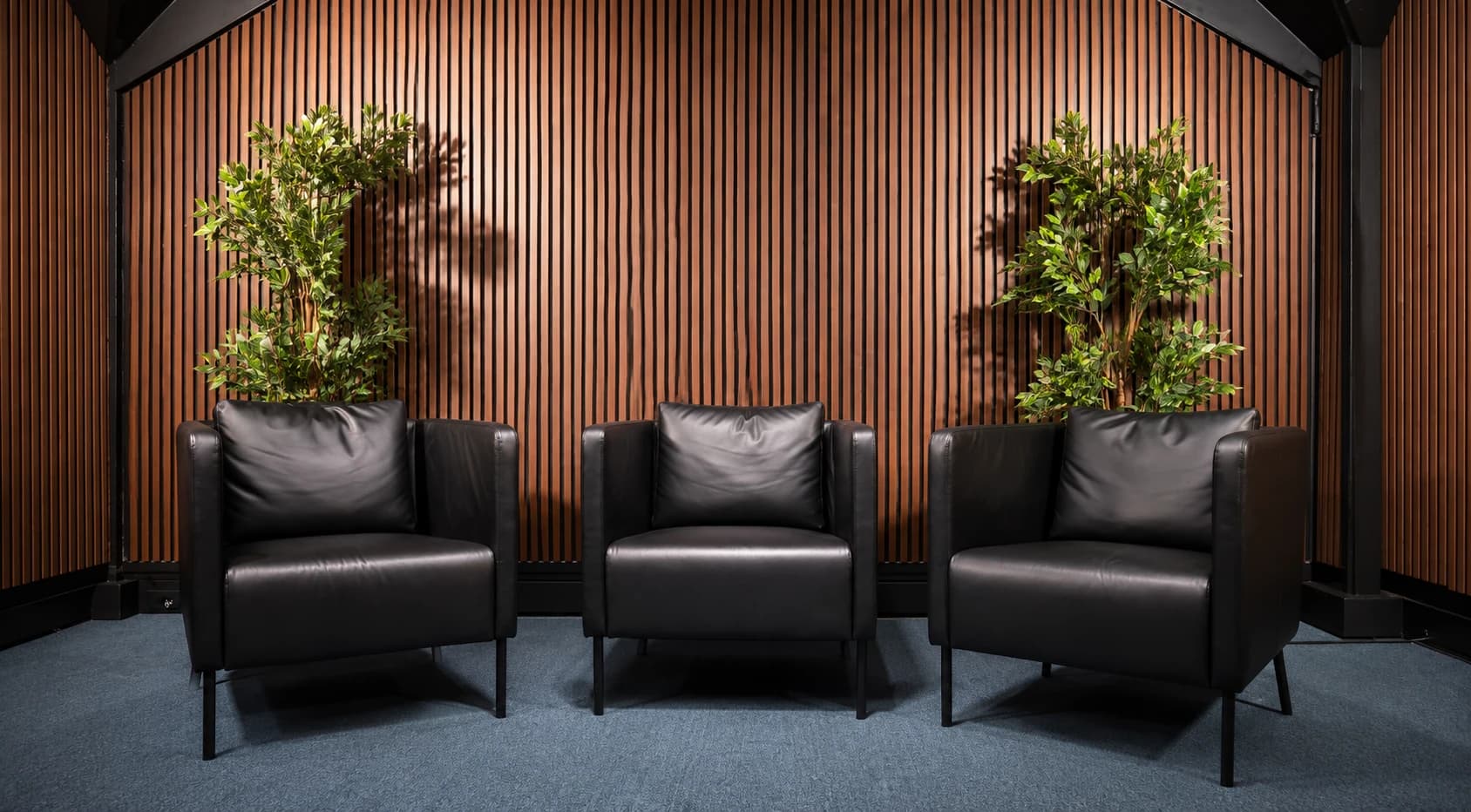 Three black leather armchairs in The Executive, without a desk, against walnut slats and plants.